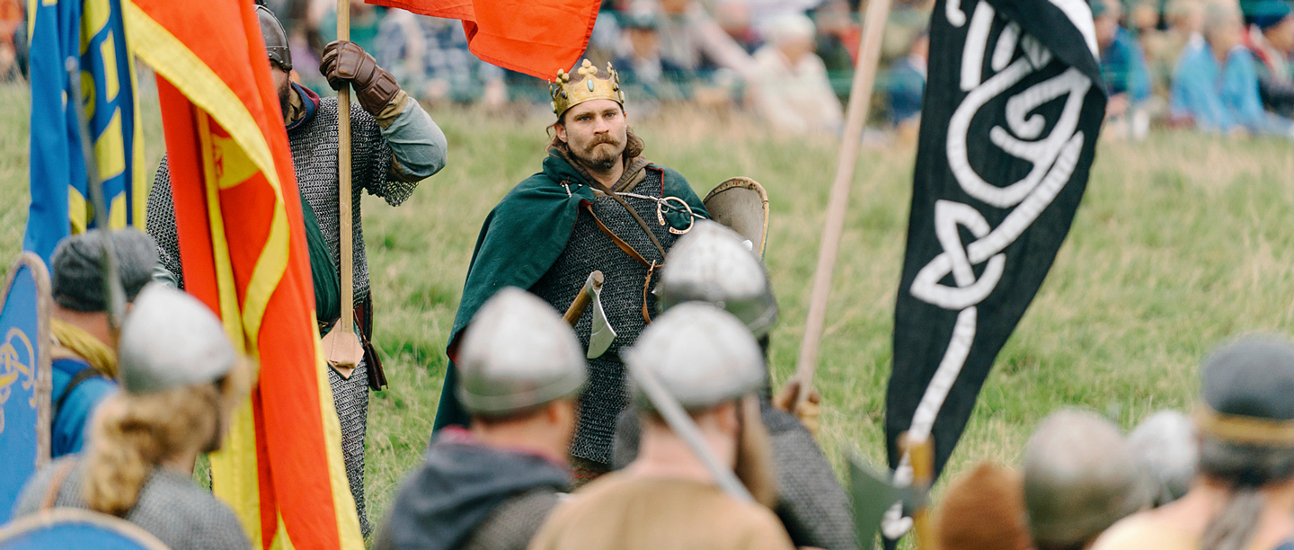 A photograph from the Battle of Hastings re-enactment event, with a man wearing a crown on horseback surrounded by men in medieval combat clothing. 