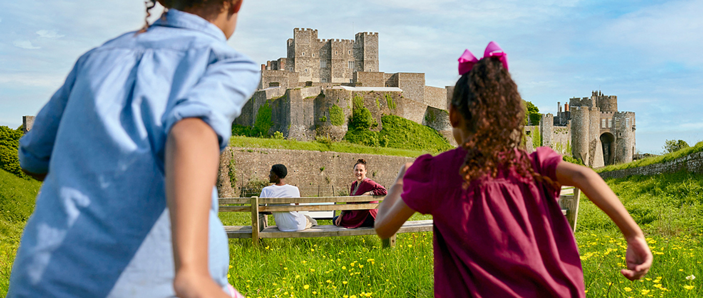 Two children run towards two adults sat on a bench outside Dover Castle.