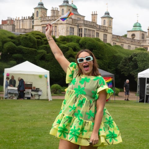 An adult wearing a colourful lime green dress and green sunglasses holds a pride flag with view of Audley End House in the background.