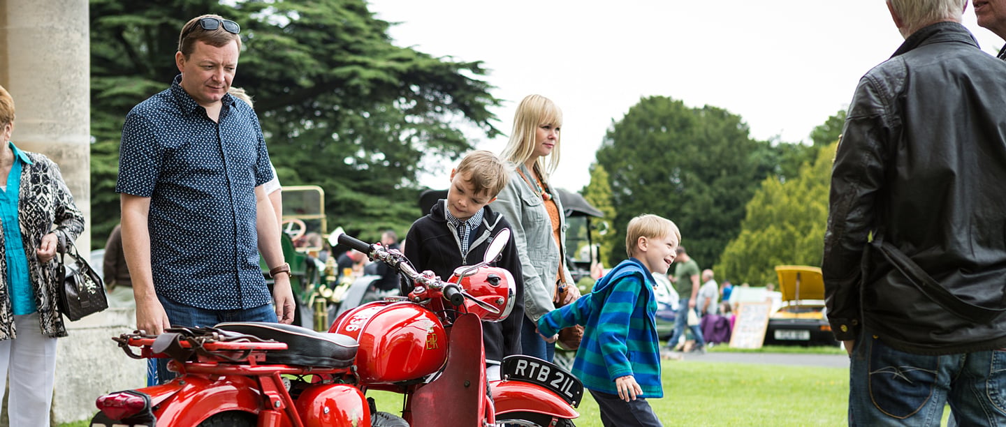 A man looks at a BSA Bantam GPO motorcycle at Brodsworth Hall Classic Car Show, surrounded by a woman and smiling children.