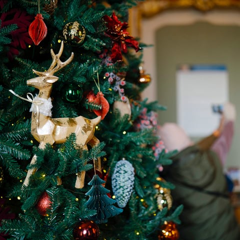 A close up view of a decorated Christmas tree with a gold reindeer. People in the background are taking a selfie.