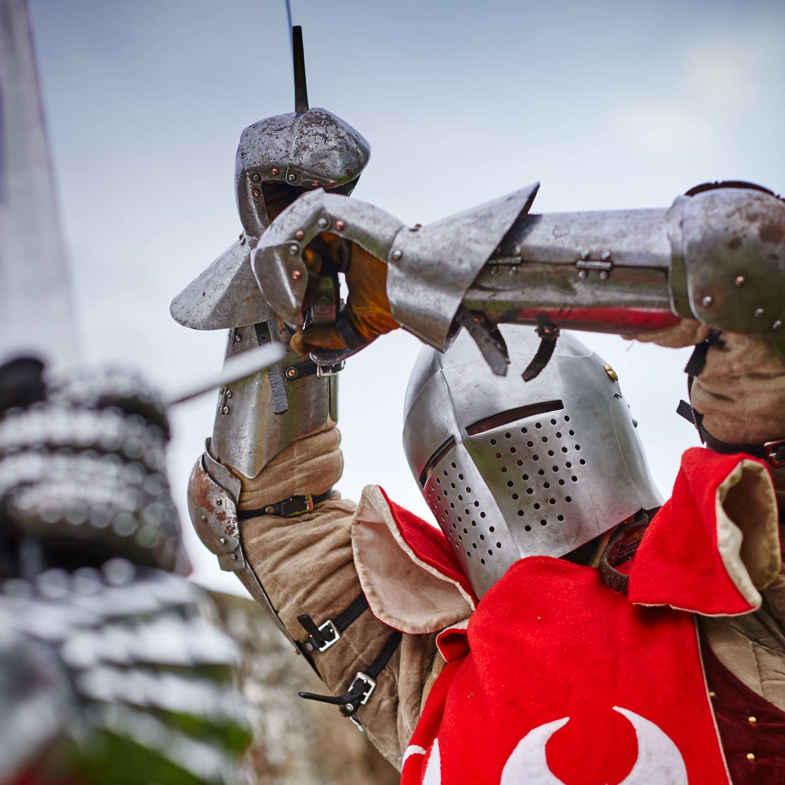 A close-up view of a reenactor dressed a Knight wears full armour and raises a sword.