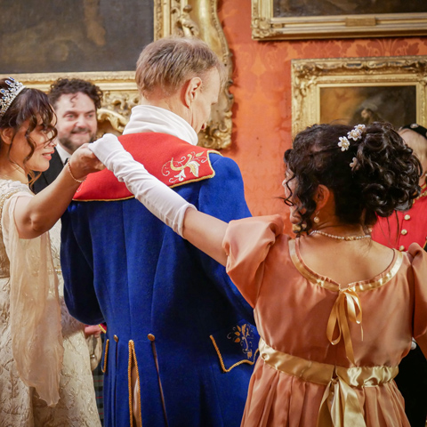 Two women extend their hands in regency dresses at a ball. 