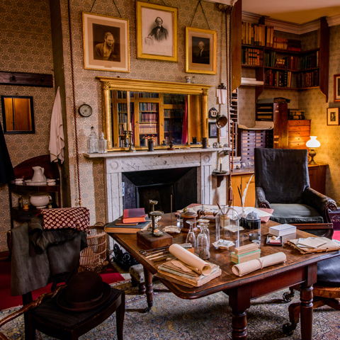 The study at Down House with portraits of Charles Darwin hung on the wall above a filled desk.
