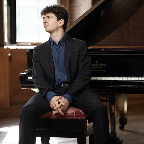 A photograph of Antoine Preat sitting in front of a grand piano on a piano stool.