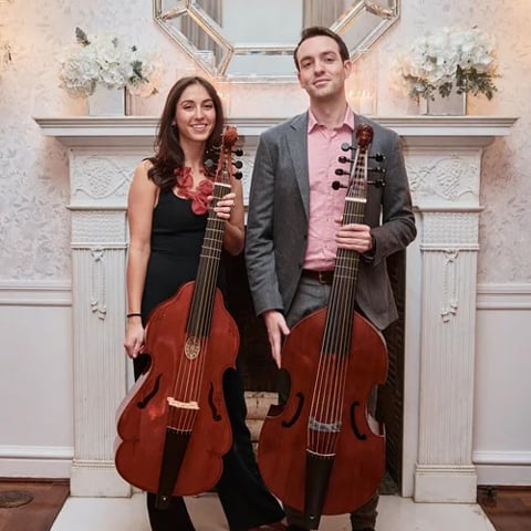 A duo hold Viol's as they stand smiling in front of a fireplace.