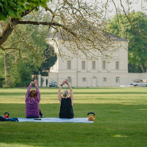Two people practice yoga poses on a yoga mat in front of Marble Hill on the lawn.