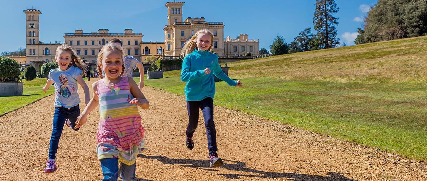 Three girls run down a gravel path while laughing in front of a large palatial house