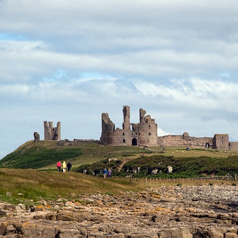 A far away view of the stone ruins at Dunstanburgh Castle on a cloudy day.