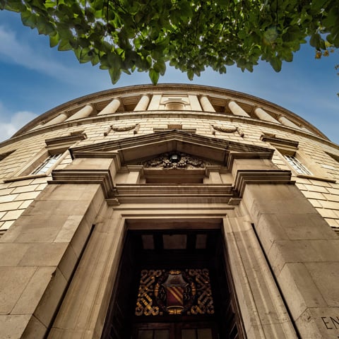 Looking up at the circular Manchester Library underneath a blue sky, with leaves on a nearby tree in view.