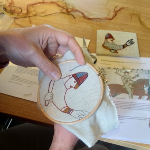 A close-up of someone doing embroidery using a fabric hoop with a Bayeux Tapestry inspired design.