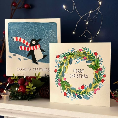 Two handmade watercolour Christmas cards stood on a white wooden fireplace.