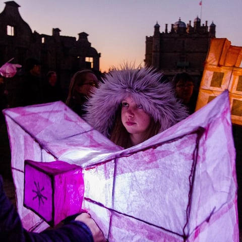 A young girl holds a lantern up in the dark in front of Bolsover Castle.