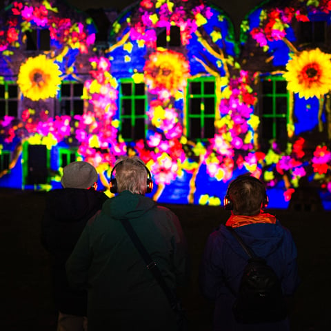 Three people stand in front of a colourful light projection display on the walls of Bolsover Castle at night.