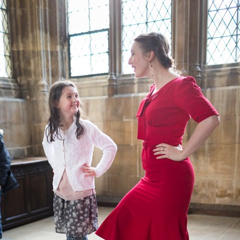 A woman wearing a red dress dances with her hands on her hips next to a young girl smiling dances with her.