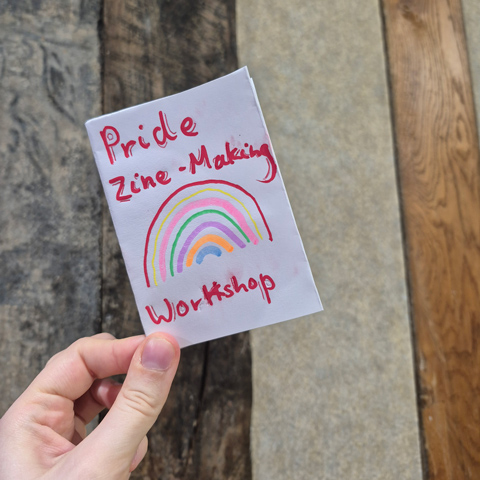 Someone holding a hand-made Zine that reads 'Pride Zine-Making Workshop' with a rainbow drawn underneath. 