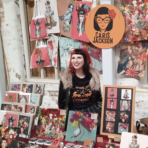 A woman smiles next to her market stall, called Caris Jackson, showcasing a variety of colourful paintings and prints.