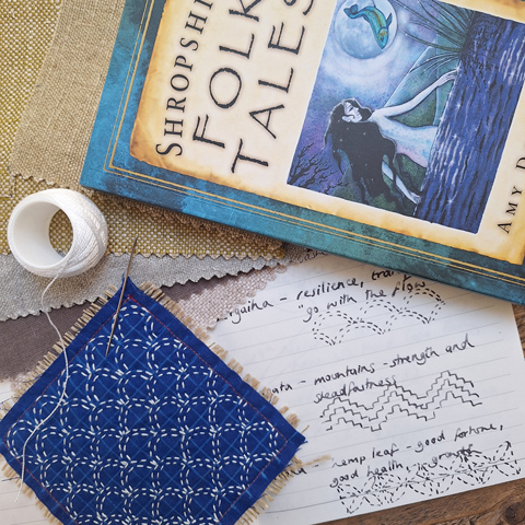 A book on Shropshire folktales laid down next to a needle and thread and a paper with illustrations on