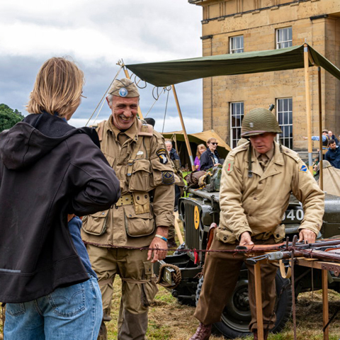 Two men wearing military Second World War uniforms showcase military objects to onlookers outside of Belsay Hall. 