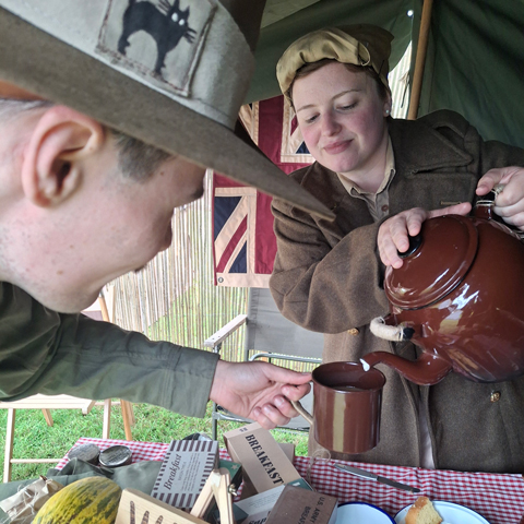A woman dressed in a 20th century style jacket serves a pot of tea.
