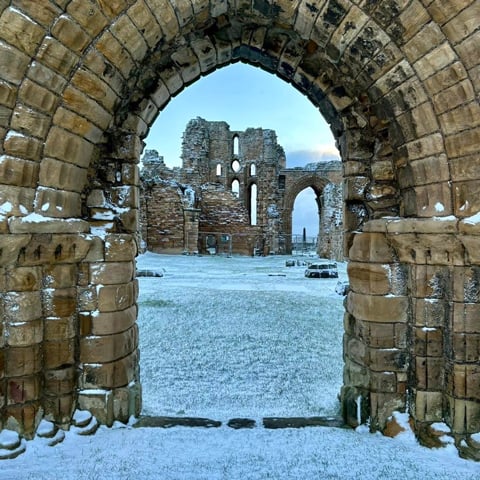 A view of Tynemouth Priory ruins covered in snow through a stone archway.