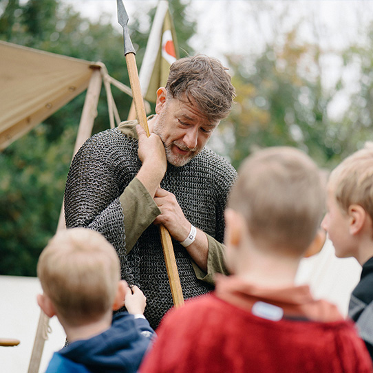 A reenactor Knight wearing chainmail stands in front of two young children. 