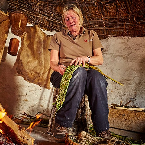 An English Heritage staff member sits on a stall in a Neolithic style hut weaving.