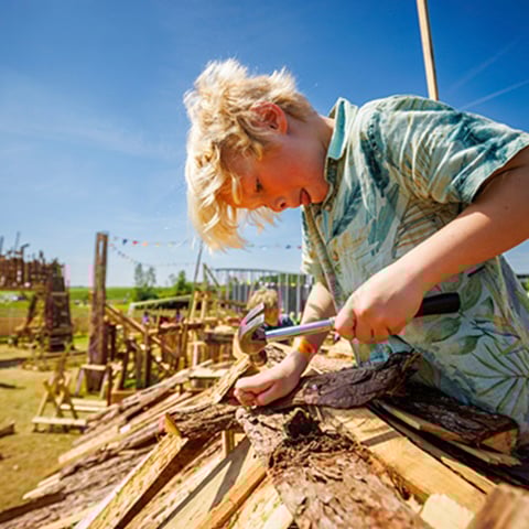 A young boy hammers a nail into a piece of wood on a sunny day.