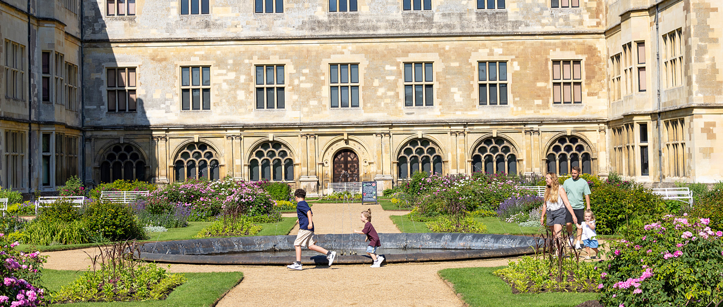 A group of adults and children wander through the gardens outside Audley End House on a sunny day.