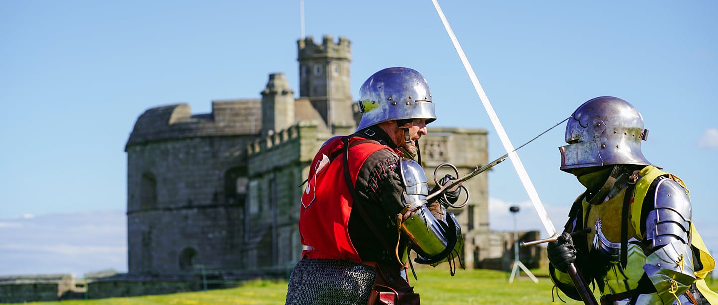 Two men dressed in Knights' armour draw their swords in front of Pendennis Castle underneath a blue sky.