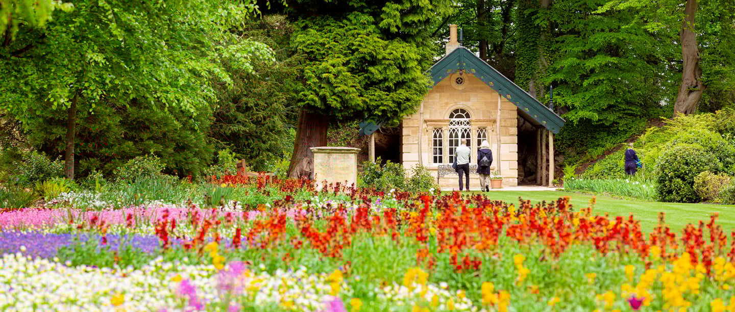 A view of the colourful flower beds in Brodsworth Hall's gardens with people walking through them next to an outbuilding in the background.