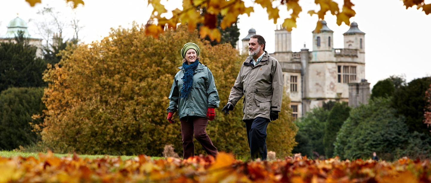 A man and a woman walk through the gardens wearing coats at Audley End in autumn.