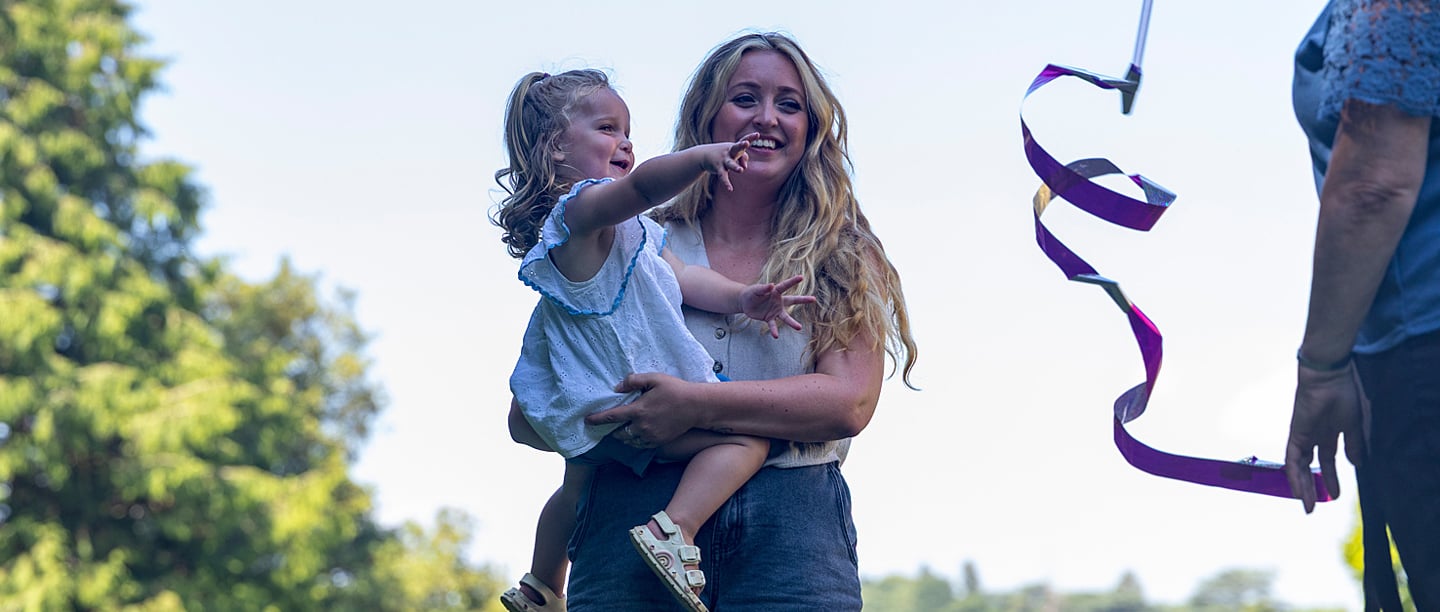 A woman smiling holds a child in her arms while someone is waving a ribbon.