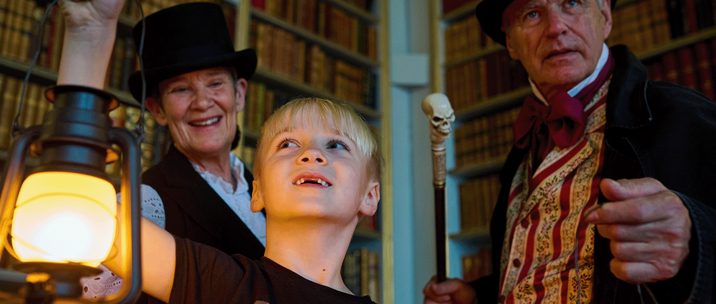 Photo of a child smiling and holding a lantern in a historic library room with two adults dressed in Victorian costume