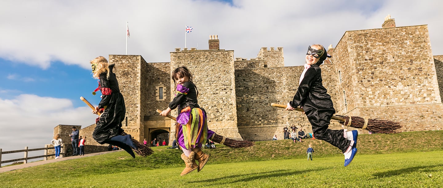 Three children dressed in Halloween costumes jump in the air holding broomsticks with Dover Castle is in the background.