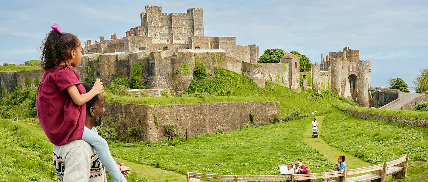 A man carries a young girl on his shoulders as they both look over at Dover Castle on a sunny day.