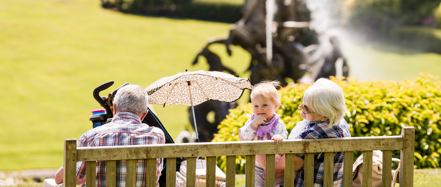 An elderly man and woman sit on a bench at Witley Court Gardens next to a child who is smiling.