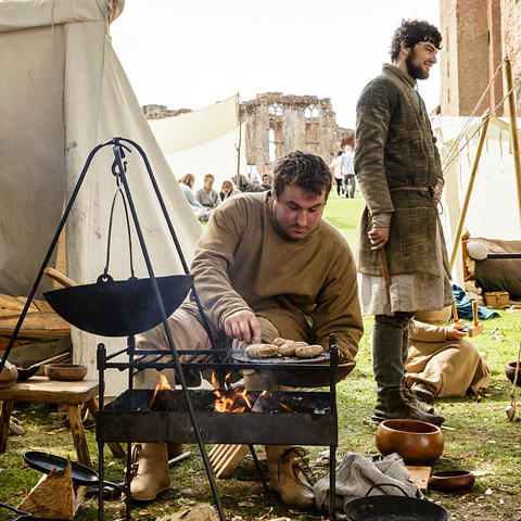 A medieval reenactor cooks food on a fire in a set up camp.